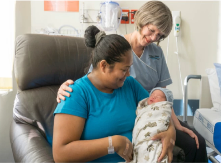 A new mother sits on a chair holding their newborn, a midwife stands to the side with their arm on the mother.