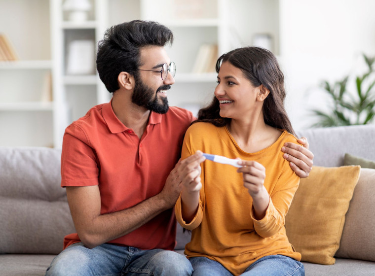 Photo of a couple sitting on a sofa smiling at each other and holding a pregnancy test stick.